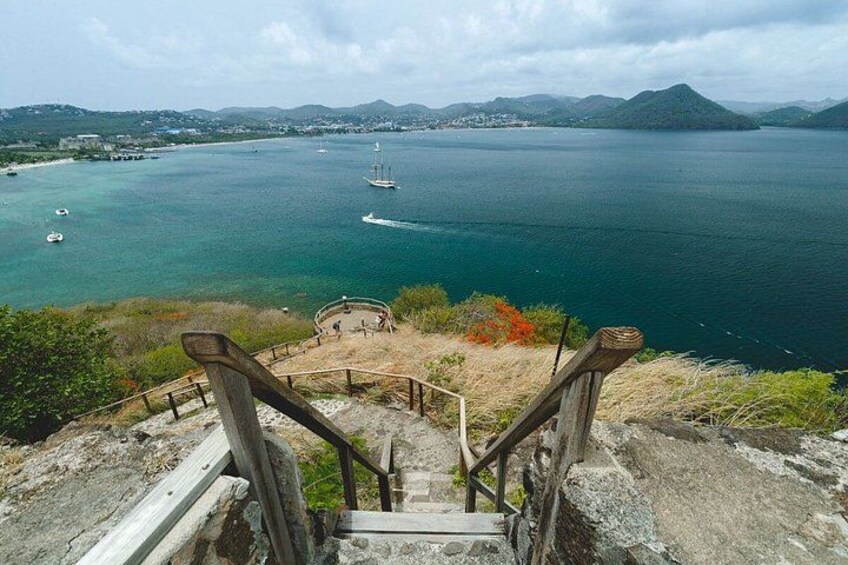 A coastal landscape with a staircase leading down to a beach, boats on the water, and distant hills. 4