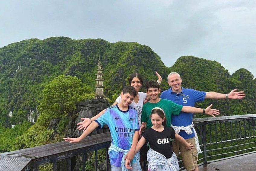 A group of people on a wooden bridge with a lush green mountain and a pagoda in the background. 2