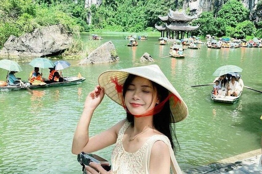 Eine Person in einem Boot mit traditionellem Hut, ein Fluss mit Booten und eine Pagode im Hintergrund. 4
