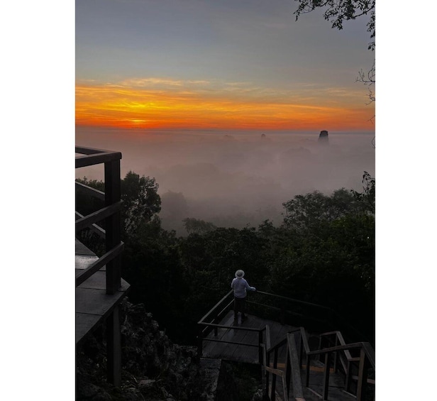 A person standing on a wooden platform overlooking a misty landscape with a sunset in the background. 1