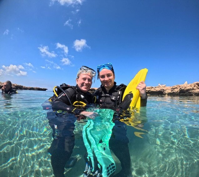 Two people in clear water with scuba gear, a yellow and a green flipper, and a rocky shoreline in the background. 2
