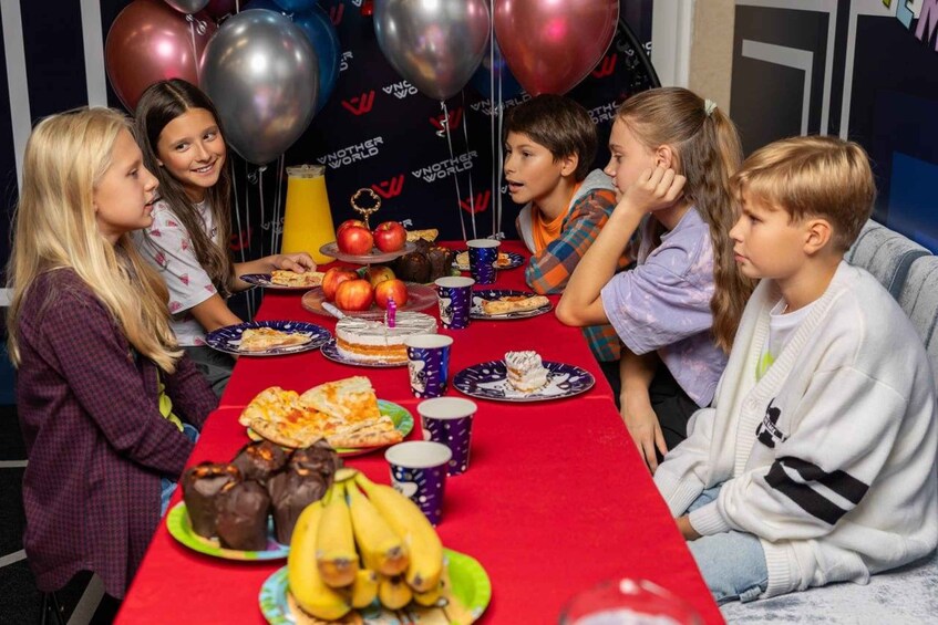 A group of children seated at a table with food and balloons in a room with a black backdrop. 3