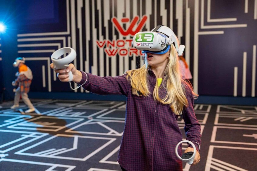 A person wearing a VR headset and holding a VR controller in a room with a geometric patterned floor and a wall with the text 'VNOT WORLDS'. 1