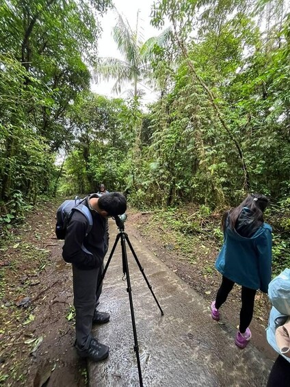 A person with a camera on a tripod, another person walking, and dense green foliage in a forested area. 3