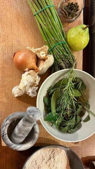 Een houten snijplank met ingrediënten voor een gerecht, waaronder knoflook, asperges, een peer, rozemarijn en een schaaltje kappertjes. 5
