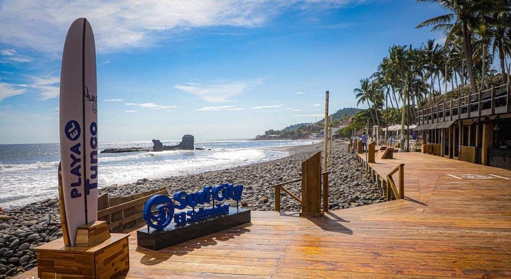 A wooden boardwalk by the beach with a surfboard and a sign that reads "PLAYA Tuxco." 1