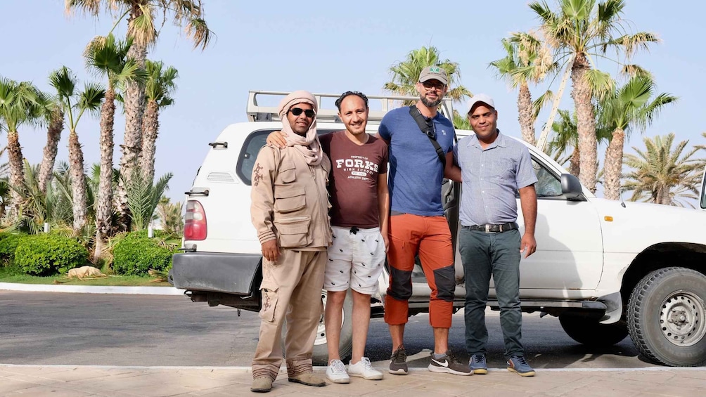 Four people standing in front of a white SUV with palm trees in the background. 3