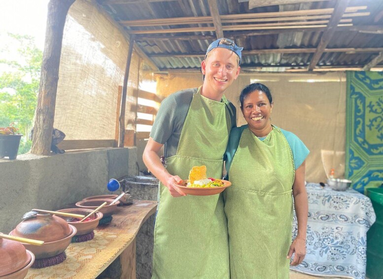 Two people in green aprons holding a plate with food, standing in a rustic outdoor kitchen with clay pots and a woven tablecloth. 2