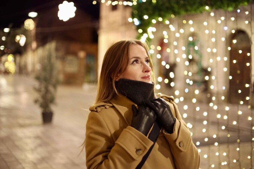 A person in a coat and gloves standing in an outdoor area with string lights and a building in the background. 5