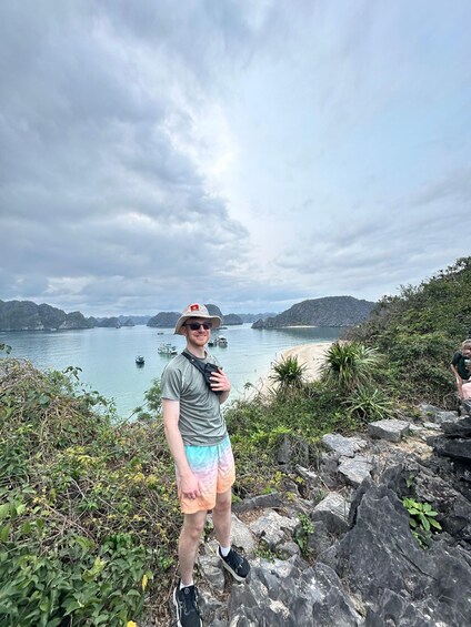 A person standing on rocks overlooking a coastal area with boats on the water and hills in the background. 3