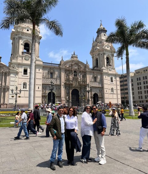 A historic building with two bell towers and a central dome, surrounded by palm trees and people. 1