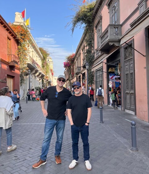 Two men standing on a cobblestone street lined with buildings, some with balconies and flowers. 4
