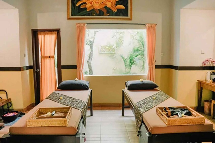 A room with two massage tables, a window with curtains, a painting, and a small shelf with items. 2