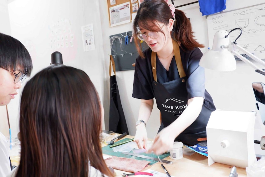 A jewelry workshop with a person working on a piece of jewelry, a white desk lamp, and a whiteboard with sketches. 1