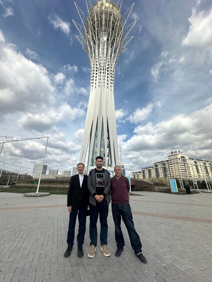 Three people standing in front of a tall tower with a unique design, under a partly cloudy sky. 4
