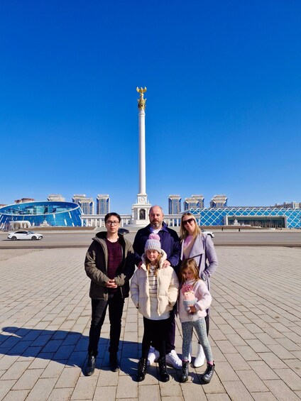A family standing in front of a tall white monument with a golden eagle on top, under a clear blue sky. 2