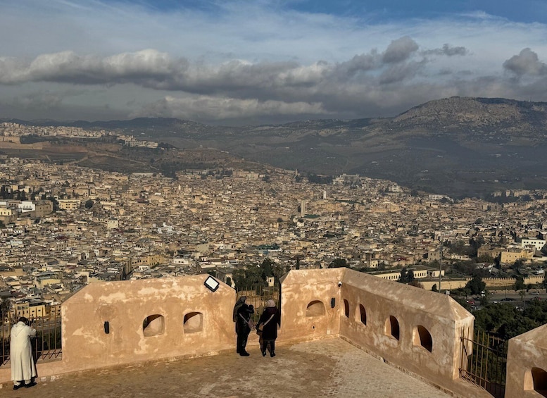 A cityscape with a view of a distant mountain range and a fortified wall with two people standing in the foreground. 4