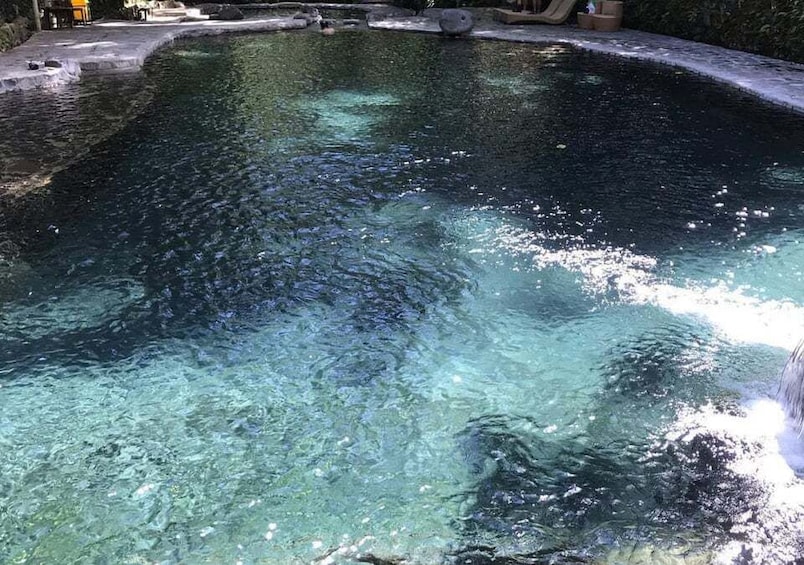 A clear swimming pool with a waterfall feature, surrounded by a stone deck and greenery. 4