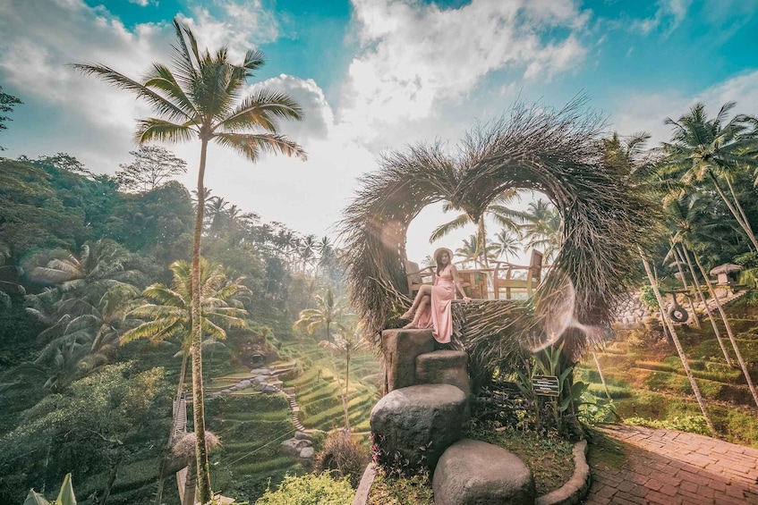 A woman in a pink dress sitting on a stone structure surrounded by lush greenery and palm trees. 5