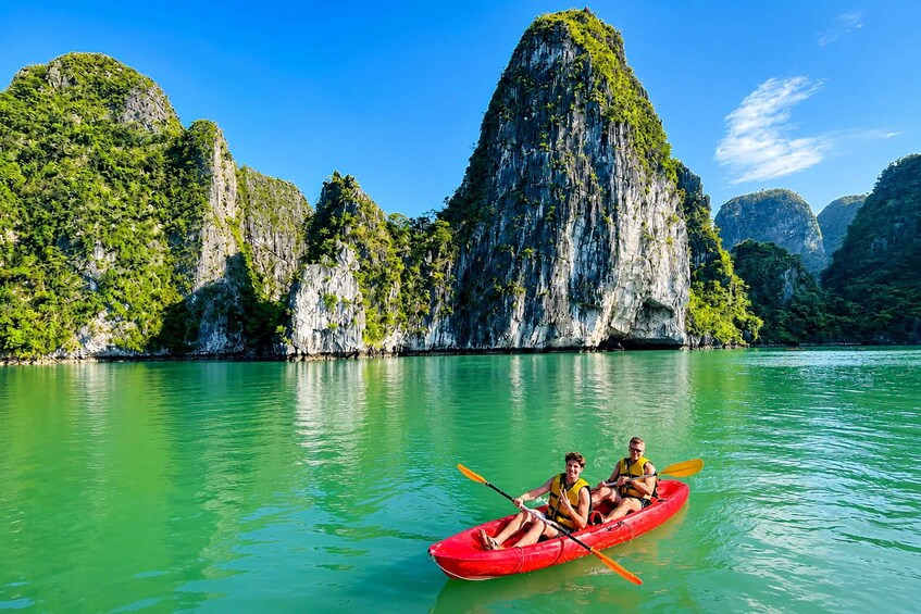 A red kayak on calm green water with two people paddling, surrounded by limestone karst formations. 4