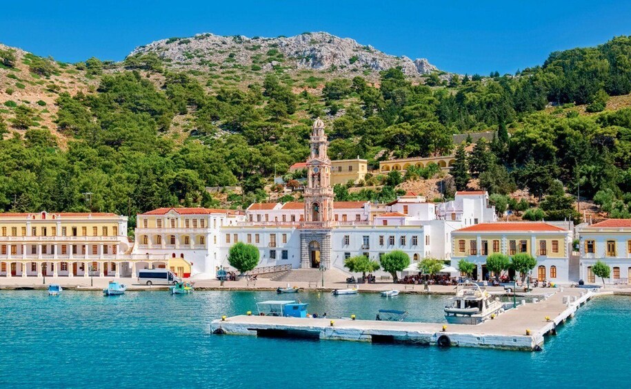 A coastal town with a prominent clock tower, white buildings with red roofs, and a dock with boats. 1