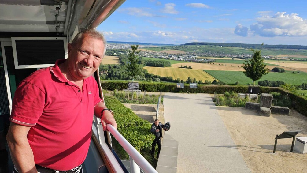 A man in a red polo shirt standing on a balcony overlooking a rural landscape with fields and a distant town. 2