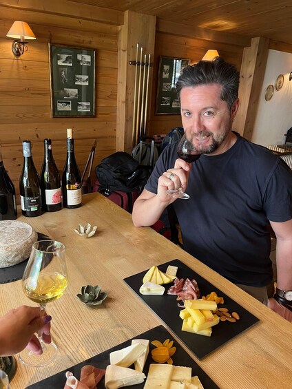 A person holding a wine glass at a wooden table with various cheeses, nuts, and bottles of wine. 2
