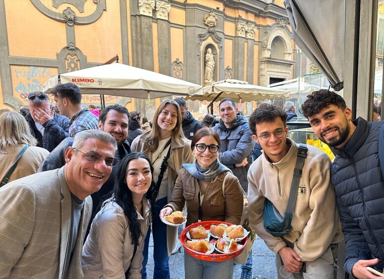 A group of people standing outside a building with a 'KIMBO' sign, holding a tray of pastries. 5