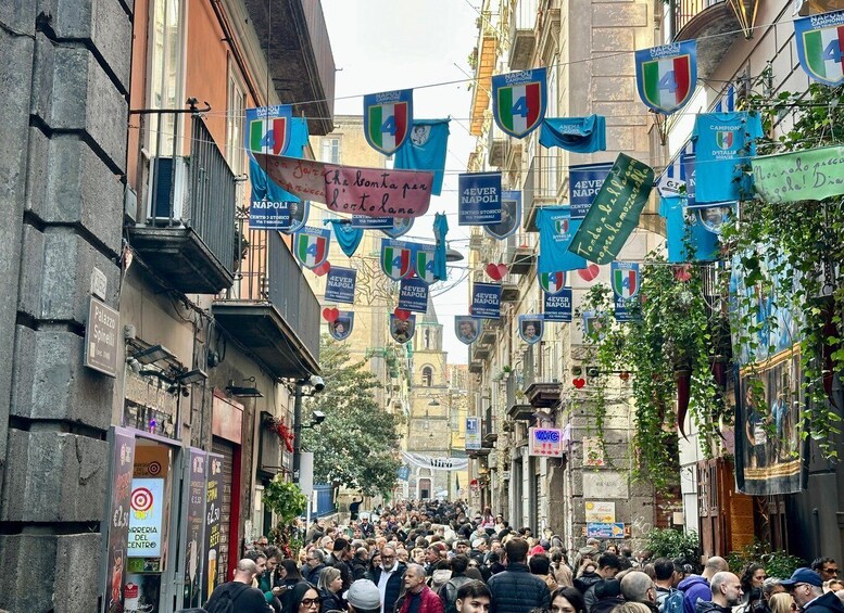A crowded street with hanging banners and signs, including 'Napoli' and '4Ever Napoli' logos. 2