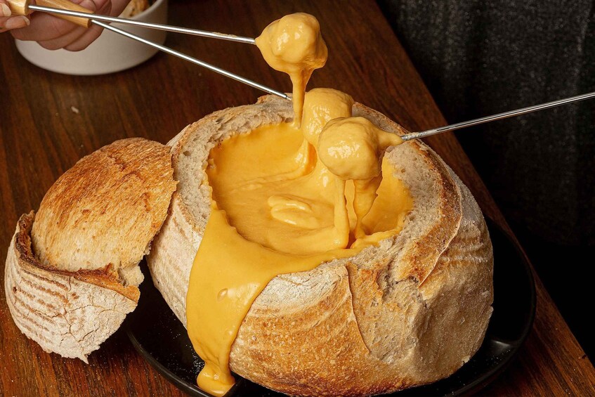 A bread bowl filled with melted cheese being held by a pair of tongs. 5