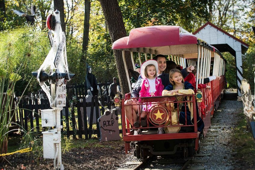 A small train with passengers, a sign with a bird, and a cemetery in the background. 1