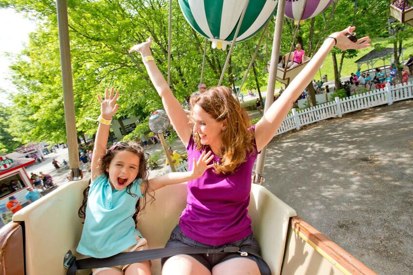 A woman and a child on a swing ride with arms raised, surrounded by trees and a white fence. 4