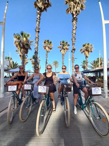 Four people on bicycles with baskets, lined up on a paved path with palm trees and a clear sky in the background. 3
