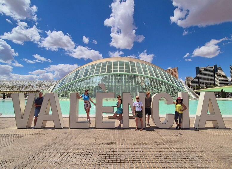 A group of people standing in front of large letters spelling 'VALENCIA' with a modern glass building in the background. 2