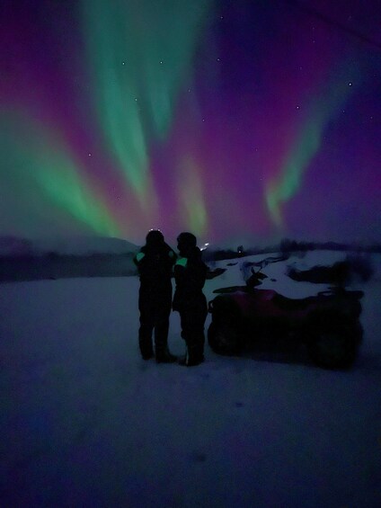 Two people standing on snow with a vehicle in the background, under a night sky with aurora borealis. 4