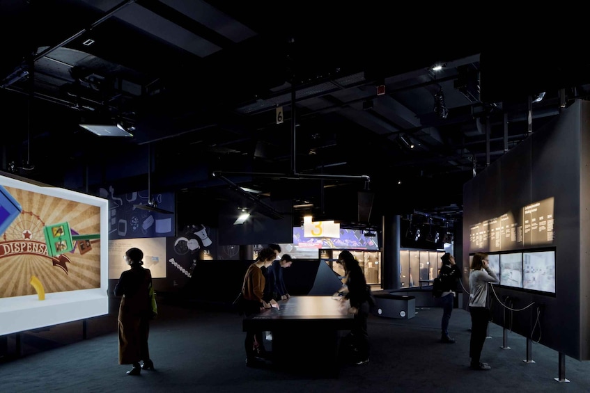 An indoor exhibition with people observing displays and a large screen with text. 2