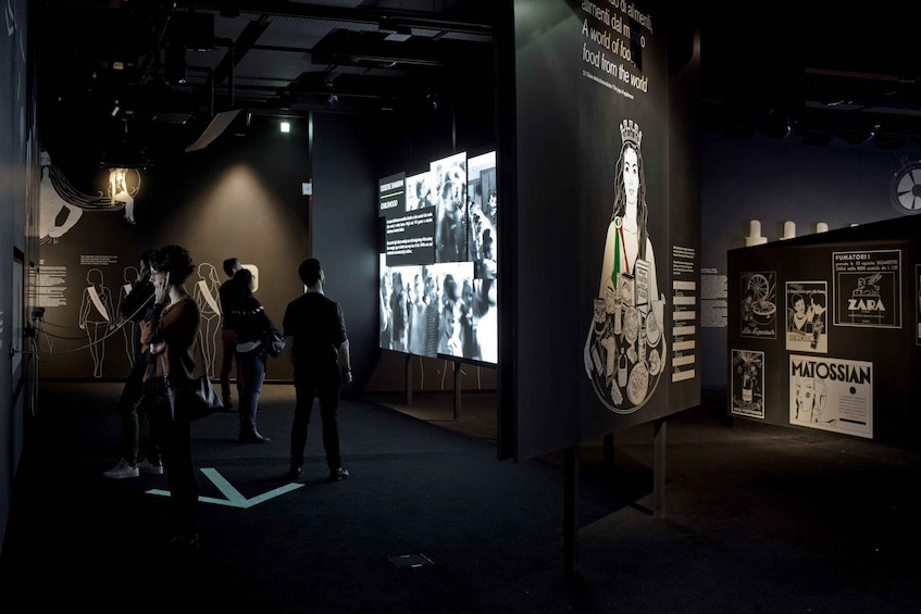 An indoor exhibition with people viewing displays, including a large screen and informational posters. 3