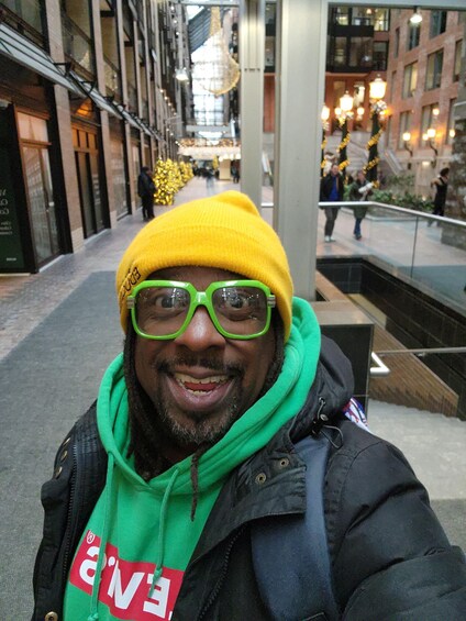 A person wearing a yellow beanie and green glasses, standing on a city street with buildings and a staircase. 5