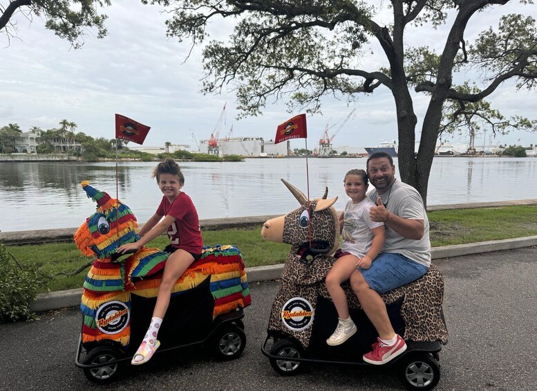 Two people riding colorful animal-shaped scooters by a body of water with construction cranes in the background. 1