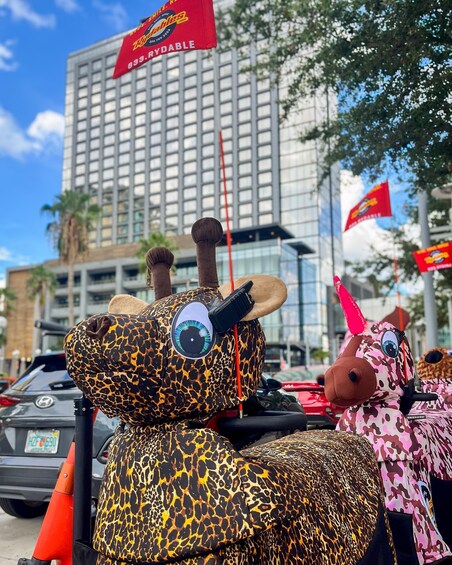 Two giraffe-shaped mascots with leopard print costumes, one with a red flag, parked on a city street with buildings and palm trees in the background. 3