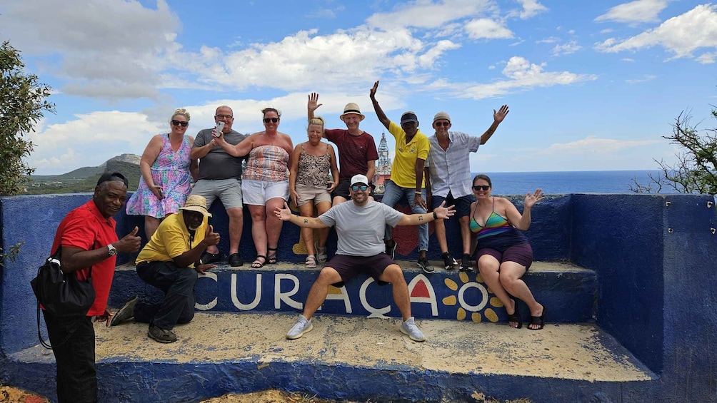 A group of people posing on a blue wall with 'CURACAO' written on it, overlooking a coastal landscape. 2
