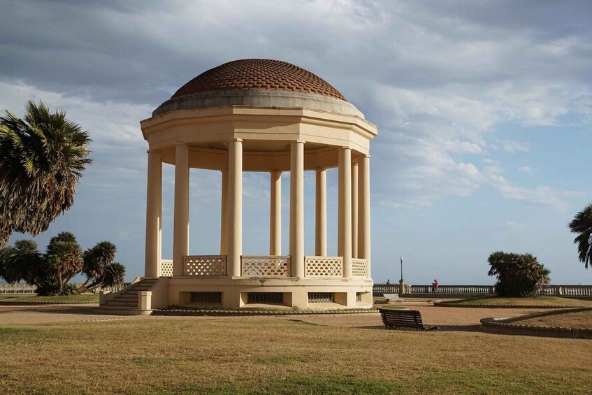 A neoclassical pavilion with a dome roof, surrounded by a park with palm trees and a distant view of the sea. 2