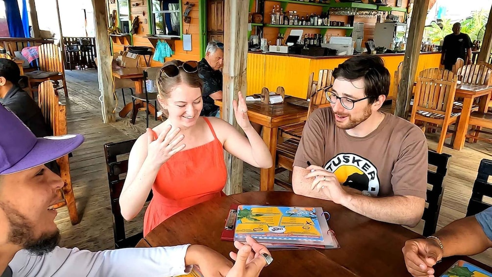 A group of people seated at a wooden table in a casual outdoor cafe with a bar in the background. 1