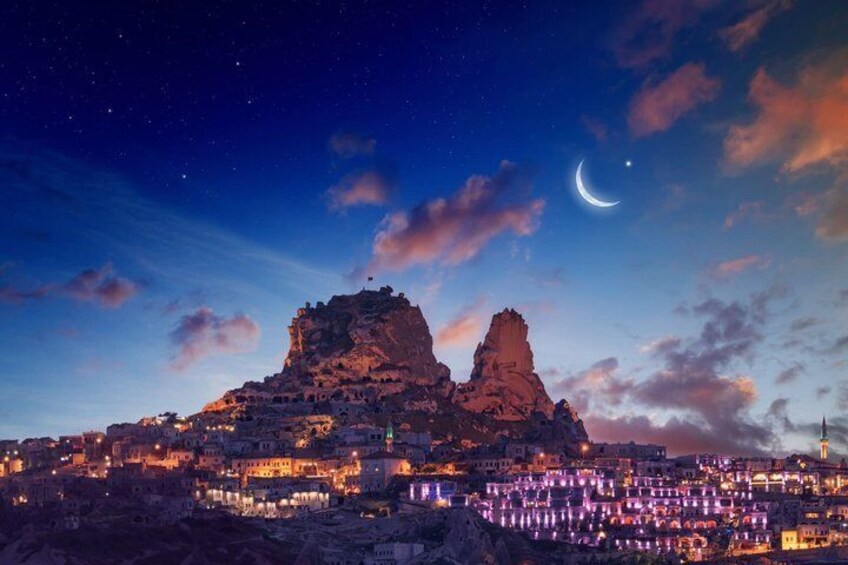 A night view of a town with illuminated buildings, a prominent rock formation, and a crescent moon in the sky. 3