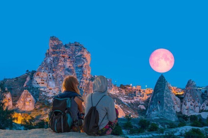 Two people sitting on a rocky outcrop, a large pink moon in the sky, and a rugged mountainous landscape. 5
