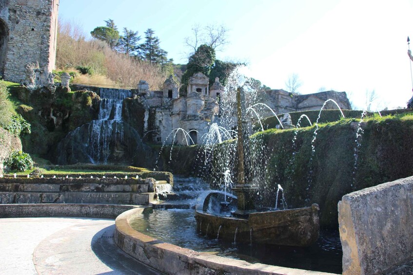 A historic stone fountain with water jets, a circular pool, and a stone bridge over a waterfall. 3