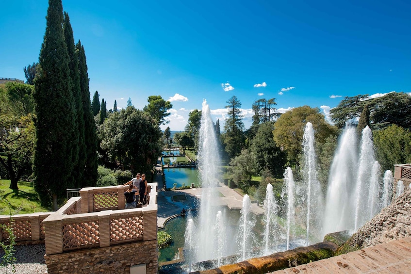 A park with a fountain, tall trees, and a clear blue sky. 1