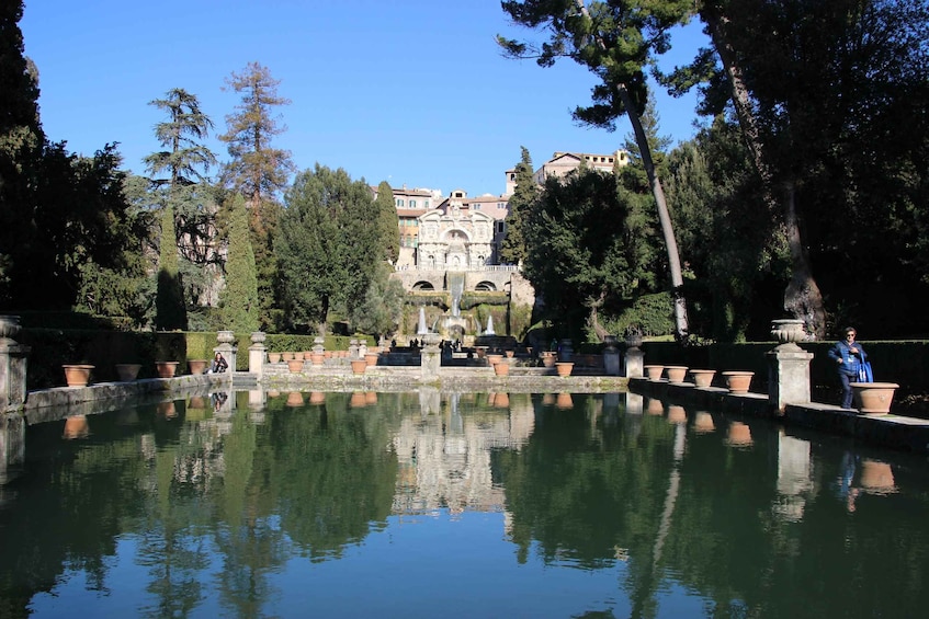 A serene garden with a reflective pond, tall trees, and a historic building with arches in the background. 4