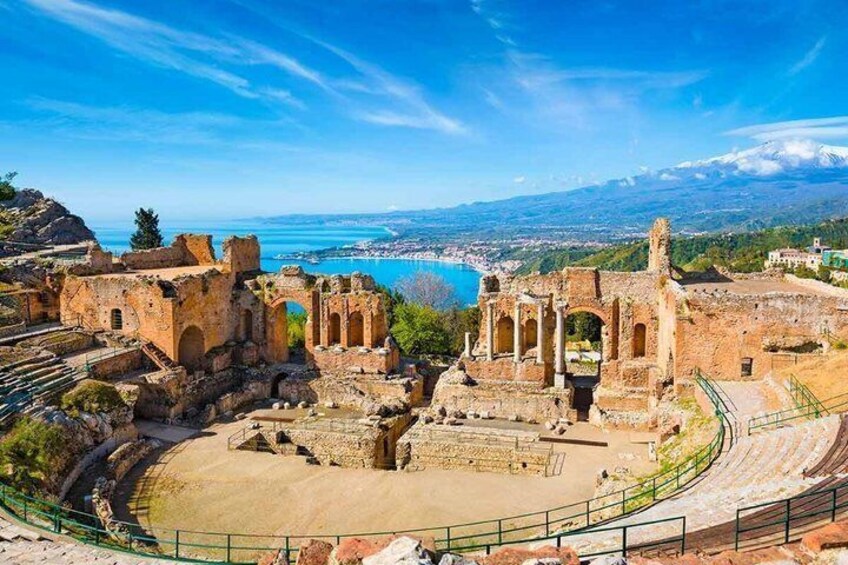 Des ruines antiques, avec un vaste amphithéâtre, un ciel d’un bleu éclatant et une vue lointaine sur une ville côtière. 1