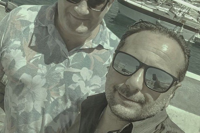 Two people wearing patterned shirts, with a body of water and boats in the background. 2